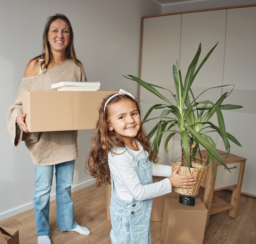 Happy mother and daughter unpacking in new house carrying moving boxes and houseplant